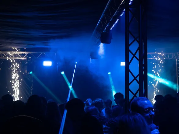 Tanzende Menschen in blauem Licht unter Traversen, grüne Lichtstrahlen und Funkenfontänen