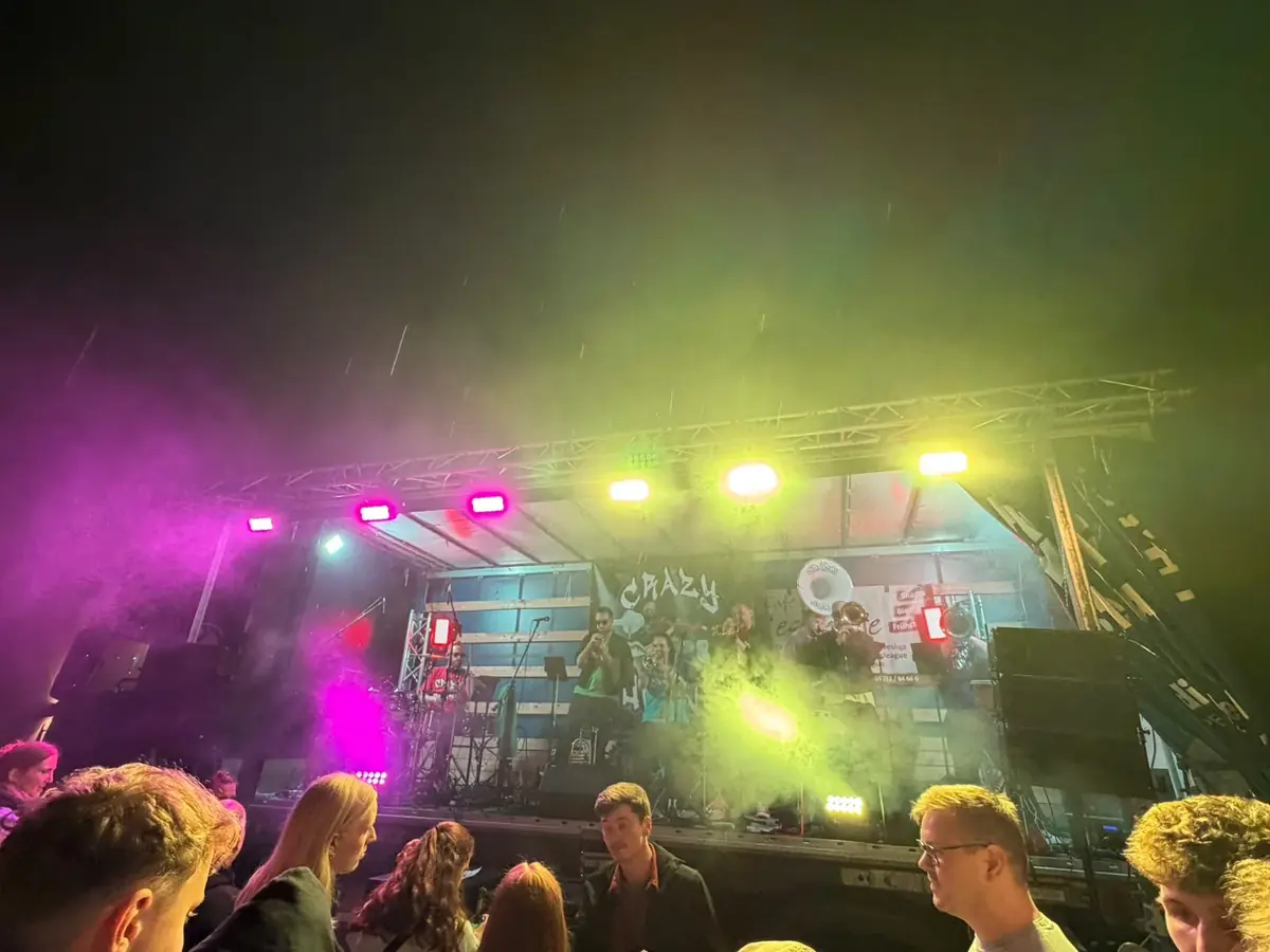 Band mit Bläsern auf einer Lkw-Bühne bei Nacht, pinkes und gelbgrünes Scheinwerferlicht im Nebel, Publikum davor