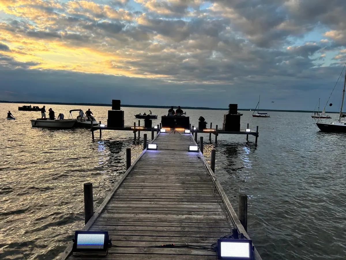 Holzsteg am See bei Sonnenuntergang mit Lautsprechern, Lichtern und DJ-Pult am Ende, Boote auf dem Wasser